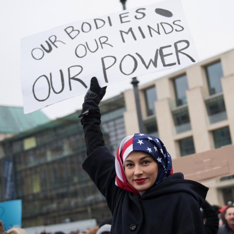 Feminist demonstrator holds a sign that says "Our bodies, our minds, our power" while wearing an American flag headscarf